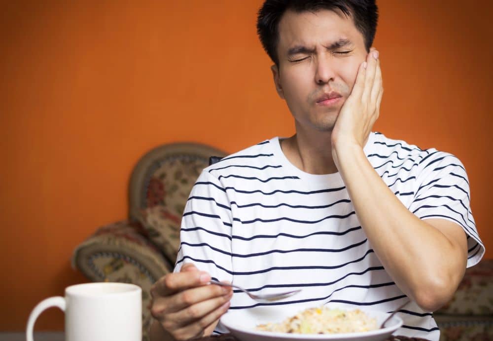 man with his left hand on his cheek in pained expression while eating